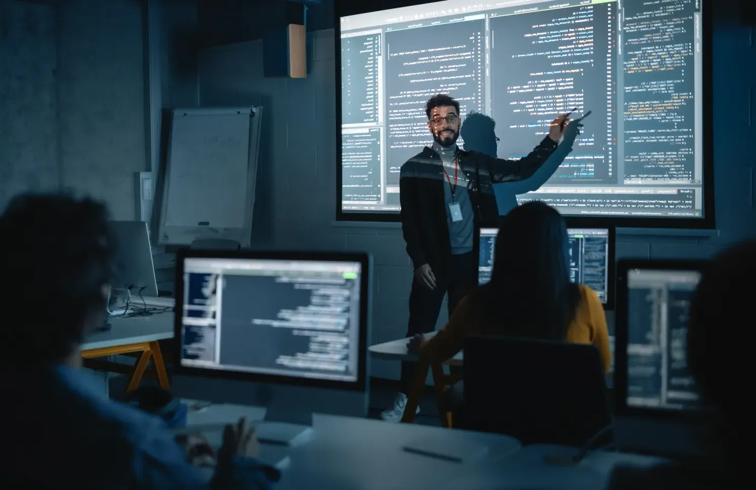 Teacher Giving Computer Science Lecture to Diverse Multiethnic Group of Female and Male Students in Dark College Room. Projecting Slideshow with Programming Code. Explaining Information Technology.